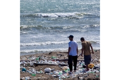 Two men on a beach surrounded by litter and plastic bottles, highlighting the issue of pollution.